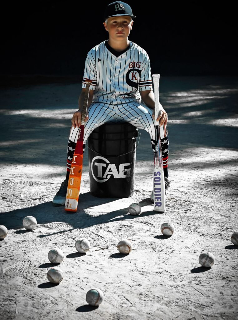 Boy in a Little League Baseball uniform, sitting on the bucket of baseballs, holding two bats.