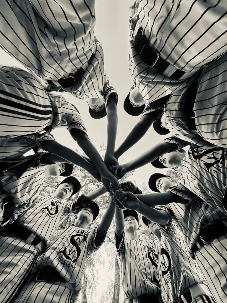 A team of Little League Baseball players extending their hands to the center of a circle. Photo taken from ground level looking up at the joined hands.