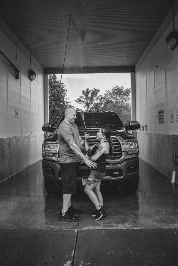 You;ng man and woman playing with the pressure washer in a carwash; Dodge pickup truck behind them in the wash bay. Black and white.