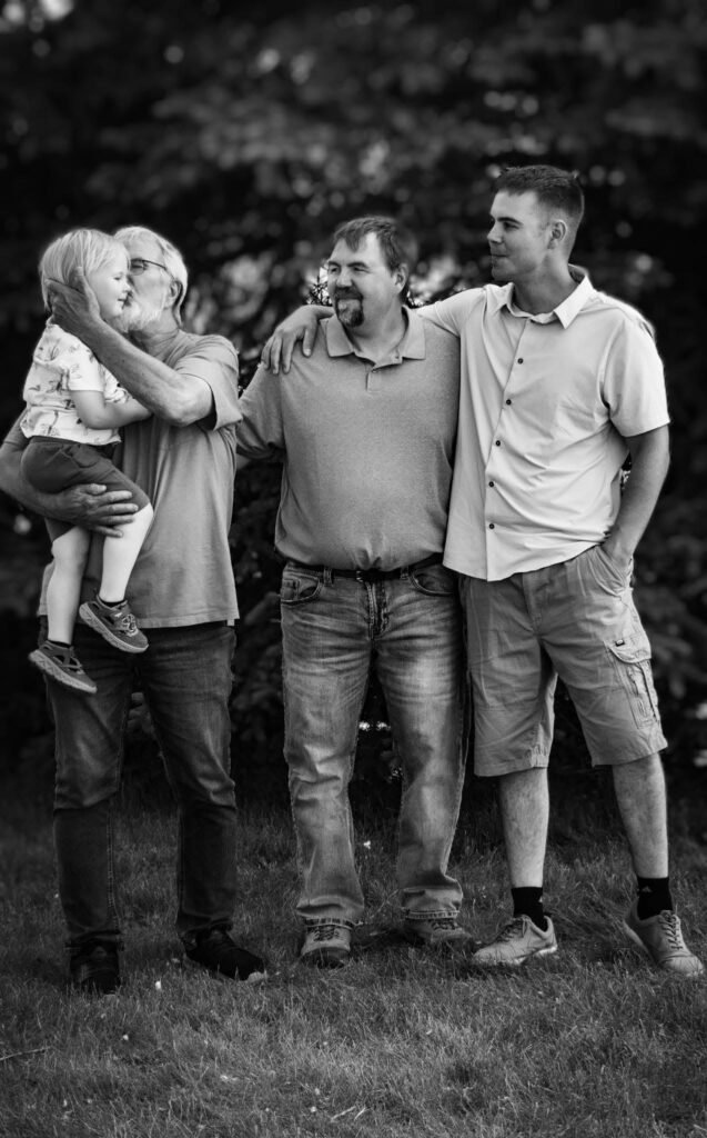 Four generations of men, great-grandfather through small child, standing in a park.