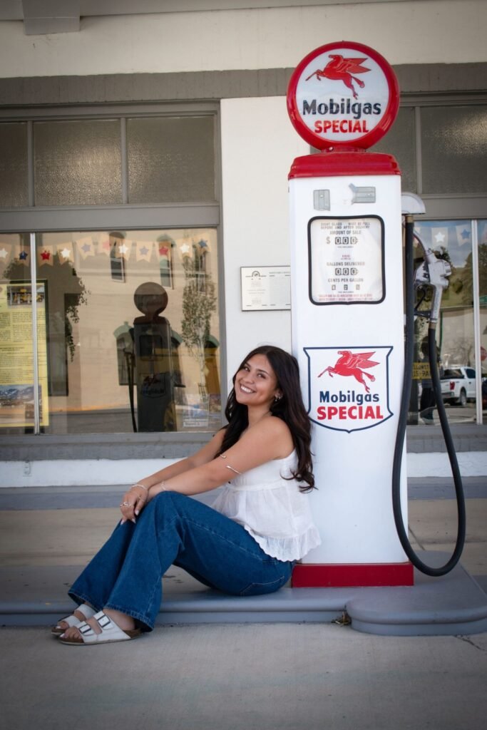 a young woman in blue jeans and a white top poses next to an antique gas pump.