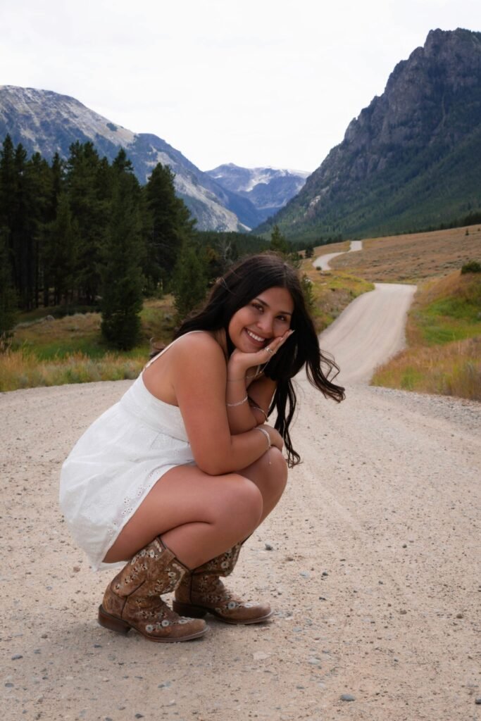 A young woman in a white dress and cowboy boots poses on a gravel road in the mountains.