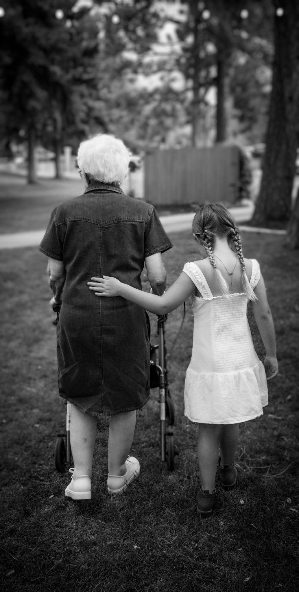 A black and white image taken from behind of an elderly lady with a walker walking through a park beside a young girl with braids in a white dress.