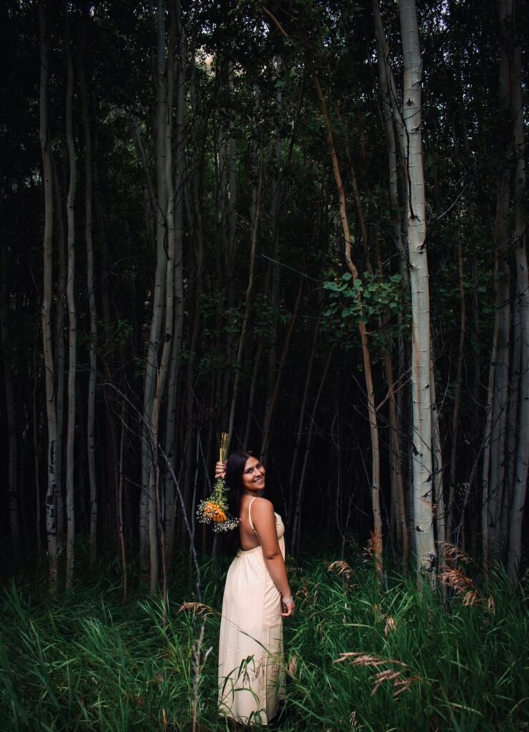 A young woman in a white dress poses with flowers in the forest.