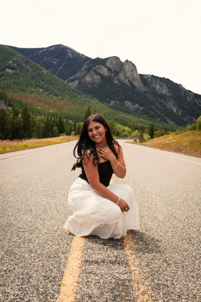 A young woman in a white skirt poses on a two-lane highway in the mountains.