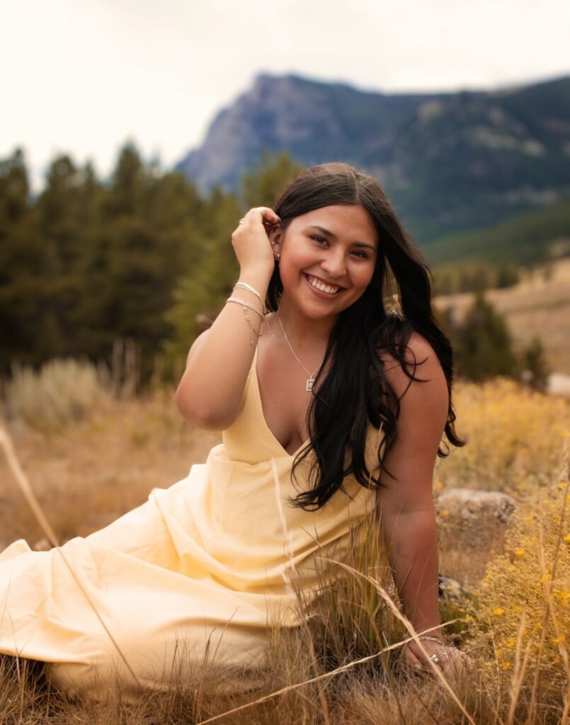 A young woman seated in the grass with evergreen trees and mountains in the background.