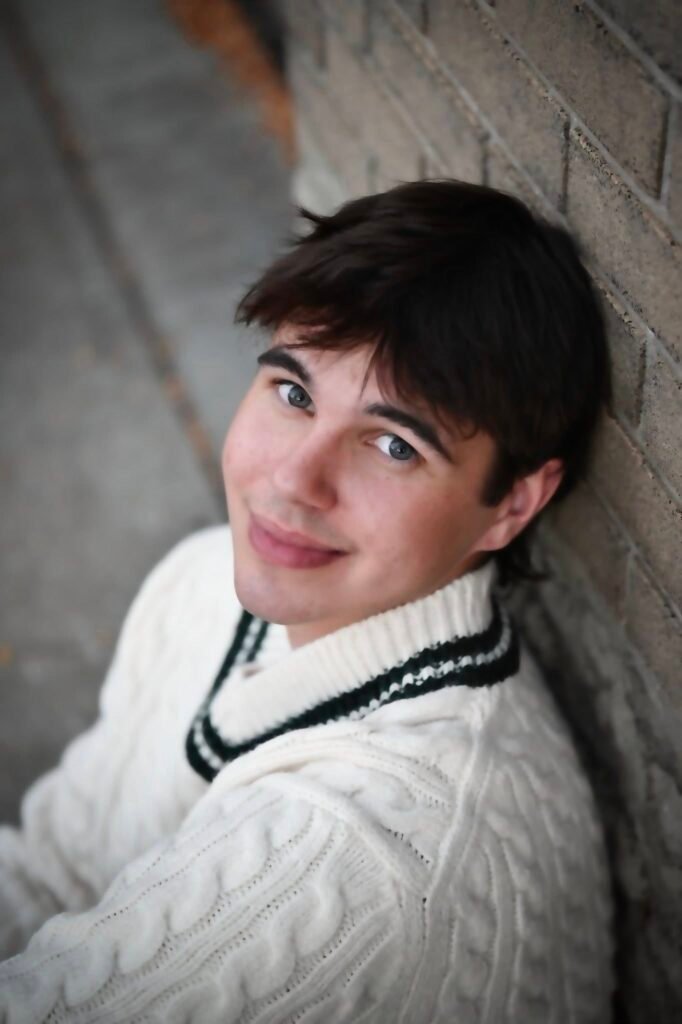 A young man in a white sweater sitting against a brick wall.