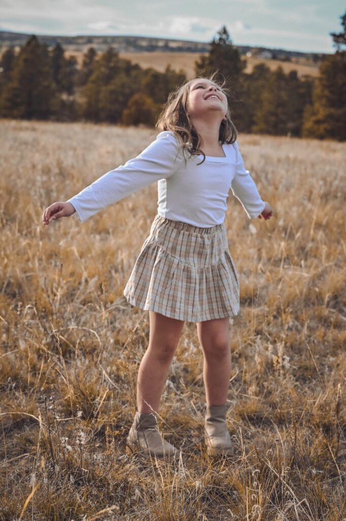 Photograph of a young girl in boots and a plaid shirt in a filed of brown grass with evergreen trees in the background.