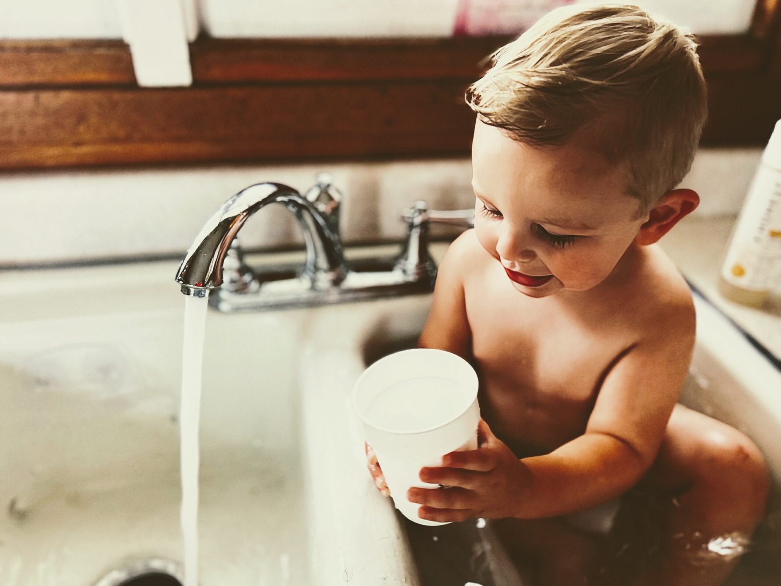 Toddler playing with a plastic cup as he takes a bath in the kitchen sink.