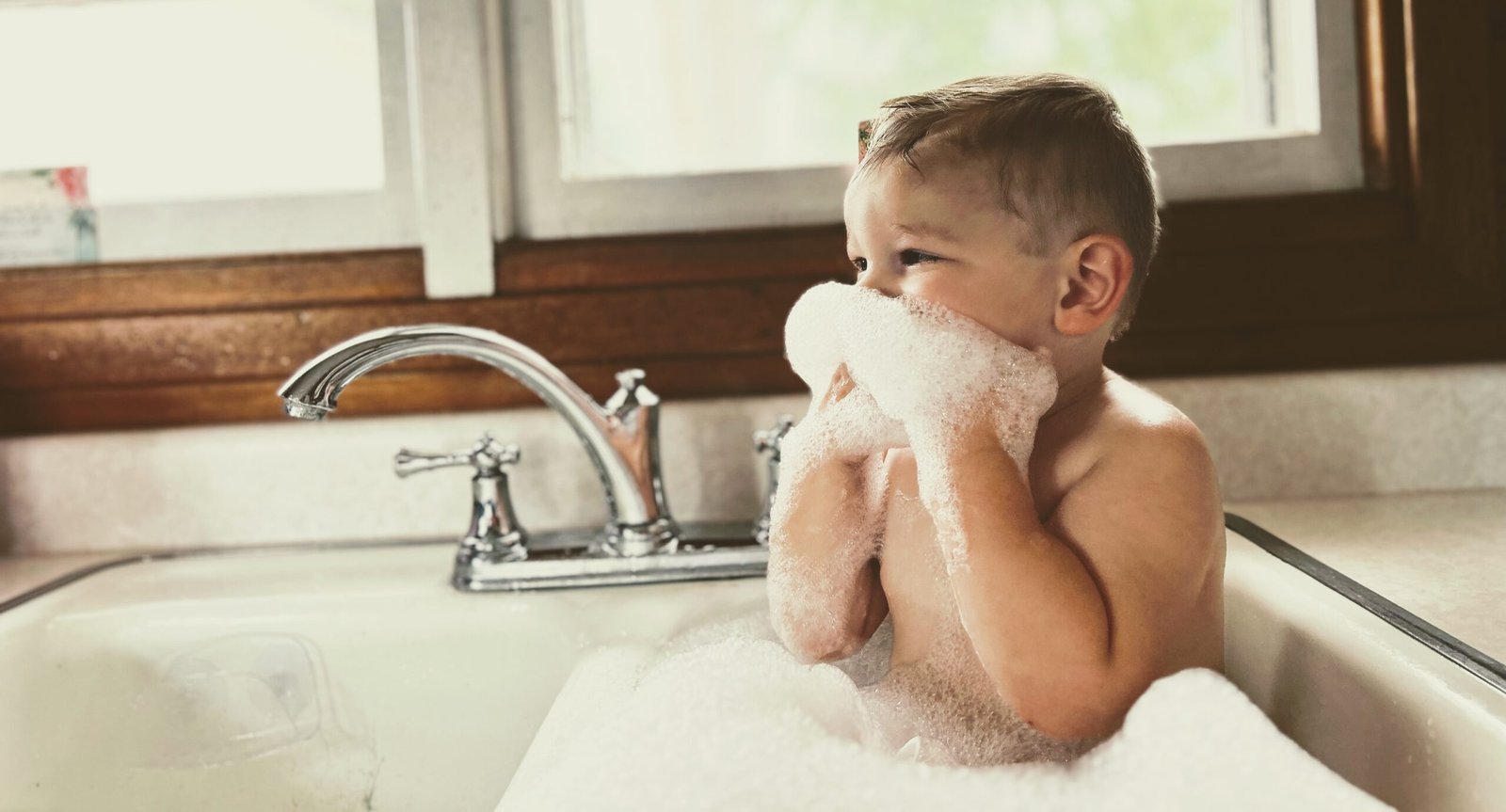 Toddler playing with suds as he takes a bath in the kitchen sink.